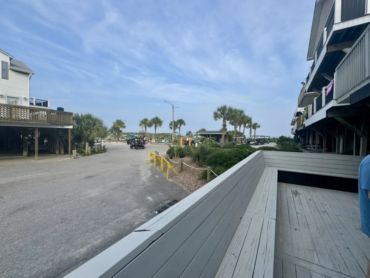 A palm-lined path leads directly to the beach, with a wooden boardwalk, nearby buildings, and the ocean visible in the distance. The scene invites guests to enjoy easy access to sun, sand, and local attractions.