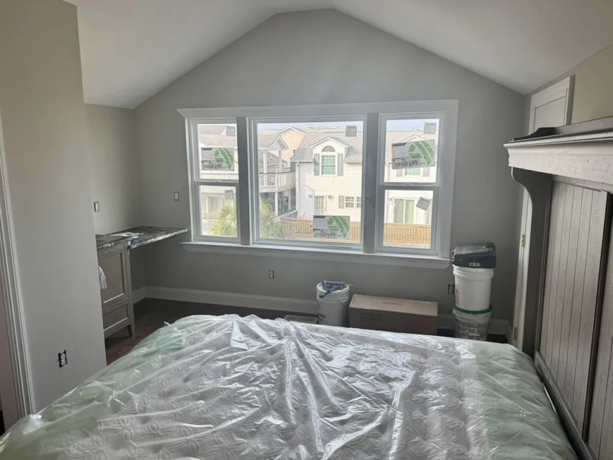 A serene bedroom with large windows letting in ample sunlight, featuring a neatly made bed with nautical-themed bedding and wooden furniture. The space feels open and bright, offering tranquil ocean views directly from the bed.