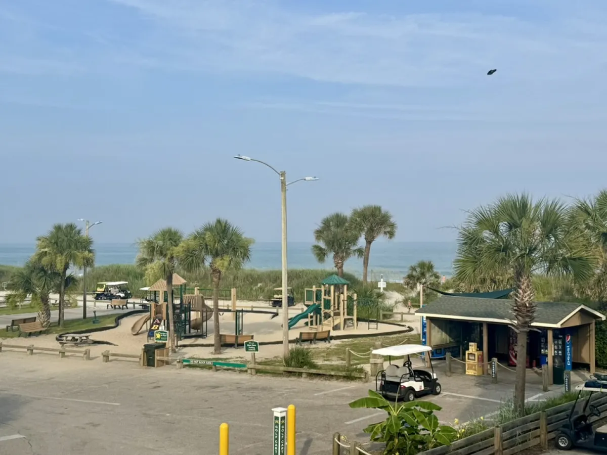 A cheerful scene featuring a playground near the beach, with a golf cart parked nearby and palm trees dotting the landscape. Children are seen playing, while families stroll and gather, enjoying the sunny, joyful atmosphere.
