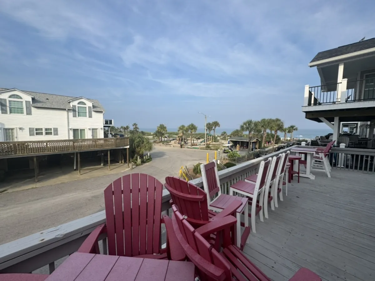This image captures a set of red and white Adirondack chairs overlooking a tranquil ocean view, framed by palm trees and neighboring houses. The scene exudes a sense of calm, inviting guests to relax and take in the natural surroundings. The spacious deck suggests private outdoor space for enjoying morning coffee or gathering with friends at sunset.