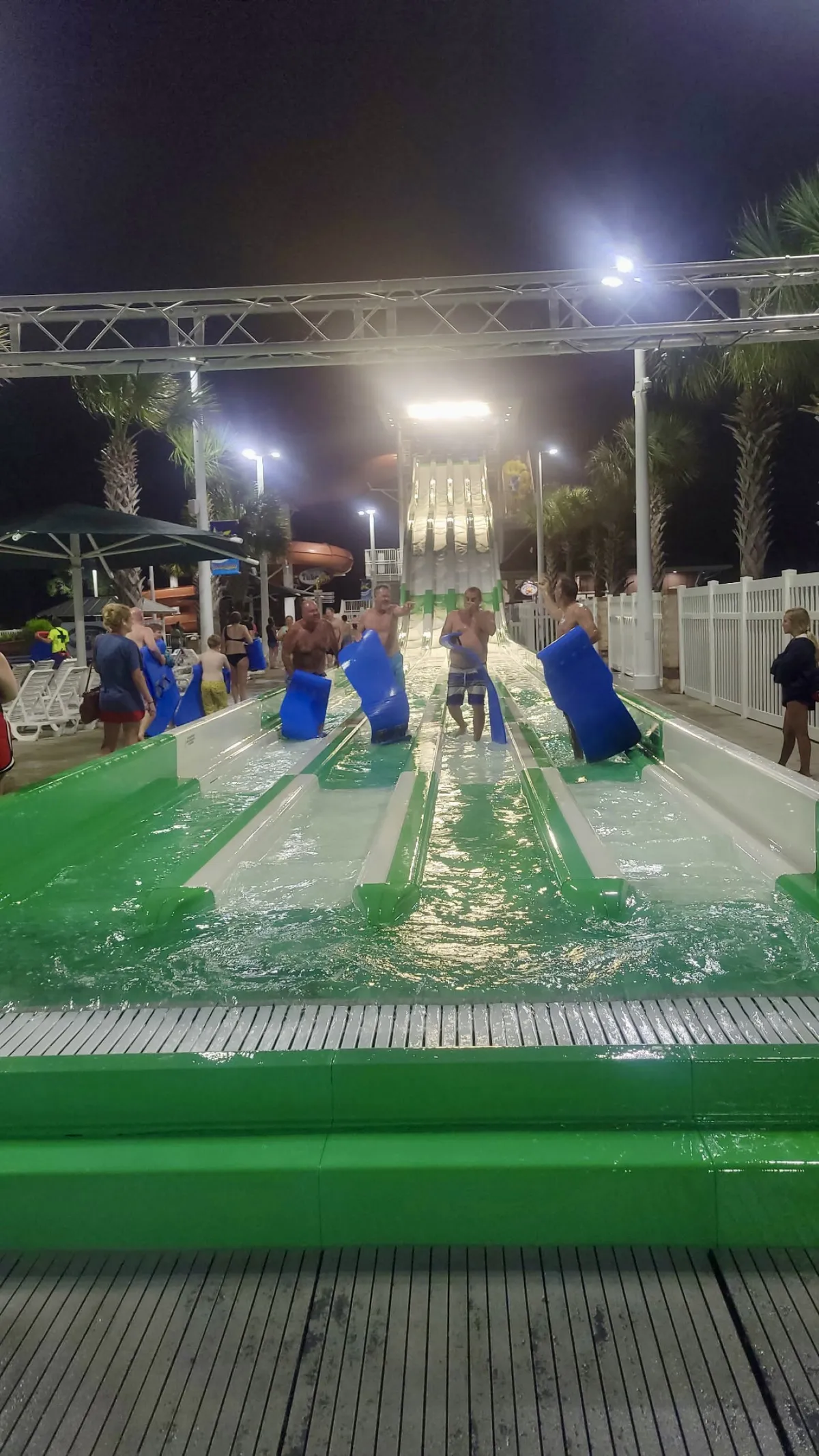 A vibrant, illuminated water park scene with colorful water slides and swimmers enjoying the pool area. Laughter and excitement fill the air as families and children take thrilling rides down the slides and gather for nighttime entertainment.