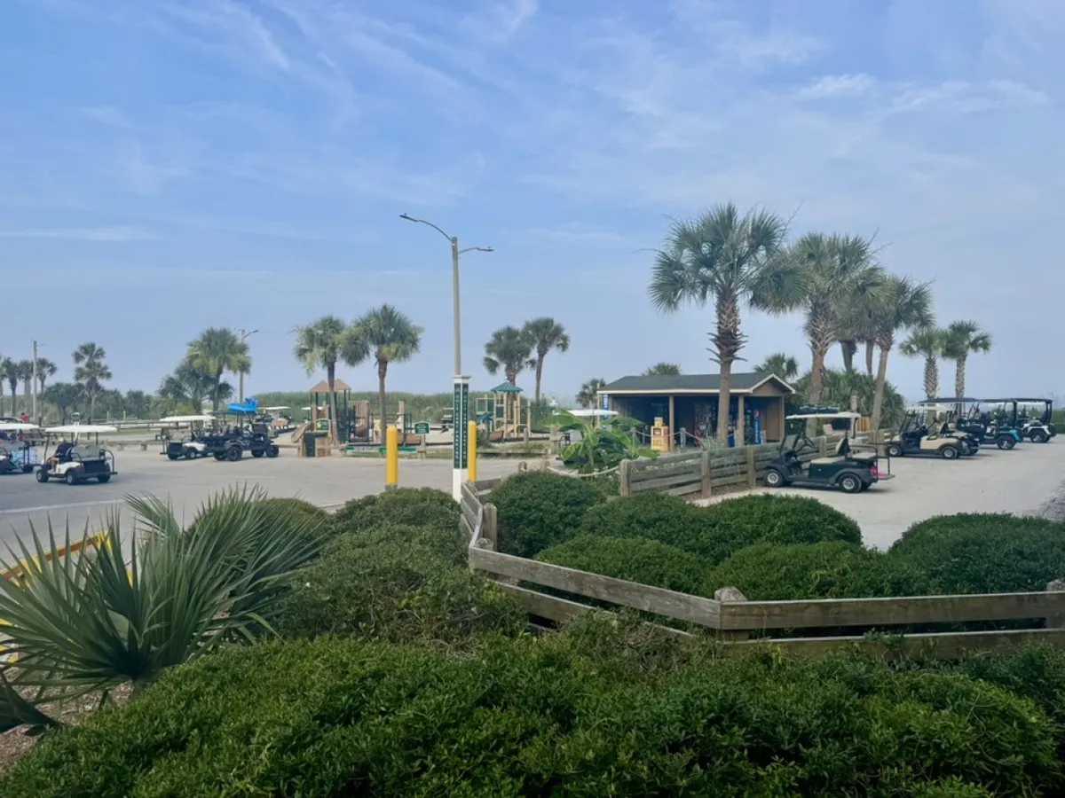A cheerful exterior scene featuring golf carts parked near palm trees, a playground, and a small building, with families enjoying the sun. The image conveys a vibrant, welcoming environment with activity and space to explore.
