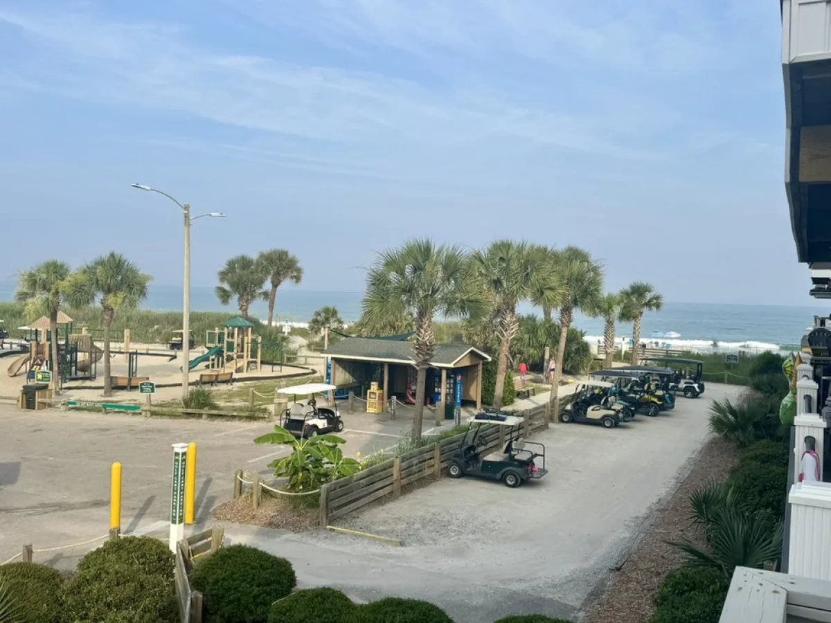 The image showcases the exterior of a luxurious, newly remodeled beach house in Myrtle Beach, South Carolina. The scene includes sweeping views of the ocean, sandy beach just steps away, palm trees swaying in the breeze, and a lively family-friendly atmosphere with children playing on a playground and golf carts ready for sunset rides.
