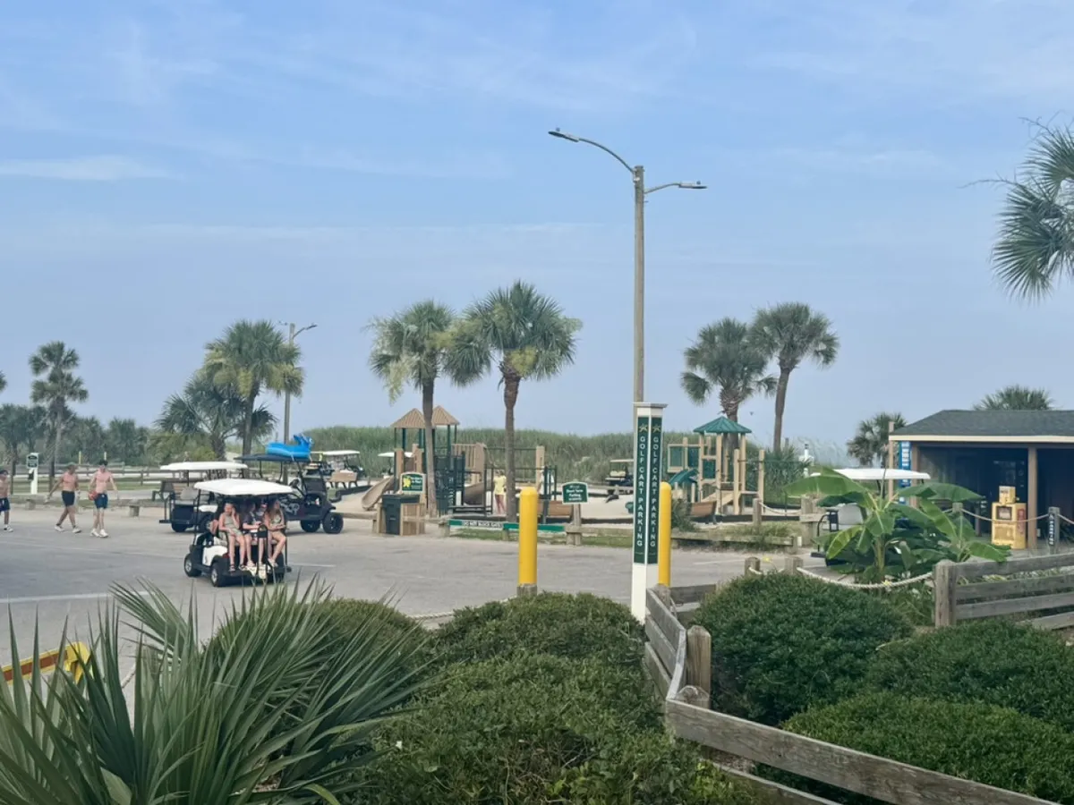 A lively scene shows palm trees framing a cheerful playground where children play, people walk along sun-dappled paths, and a golf cart is parked nearby, suggesting both fun and convenience.